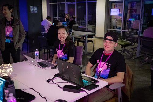 2 Space Apps participants in front of laptops in a purple and pink light setting